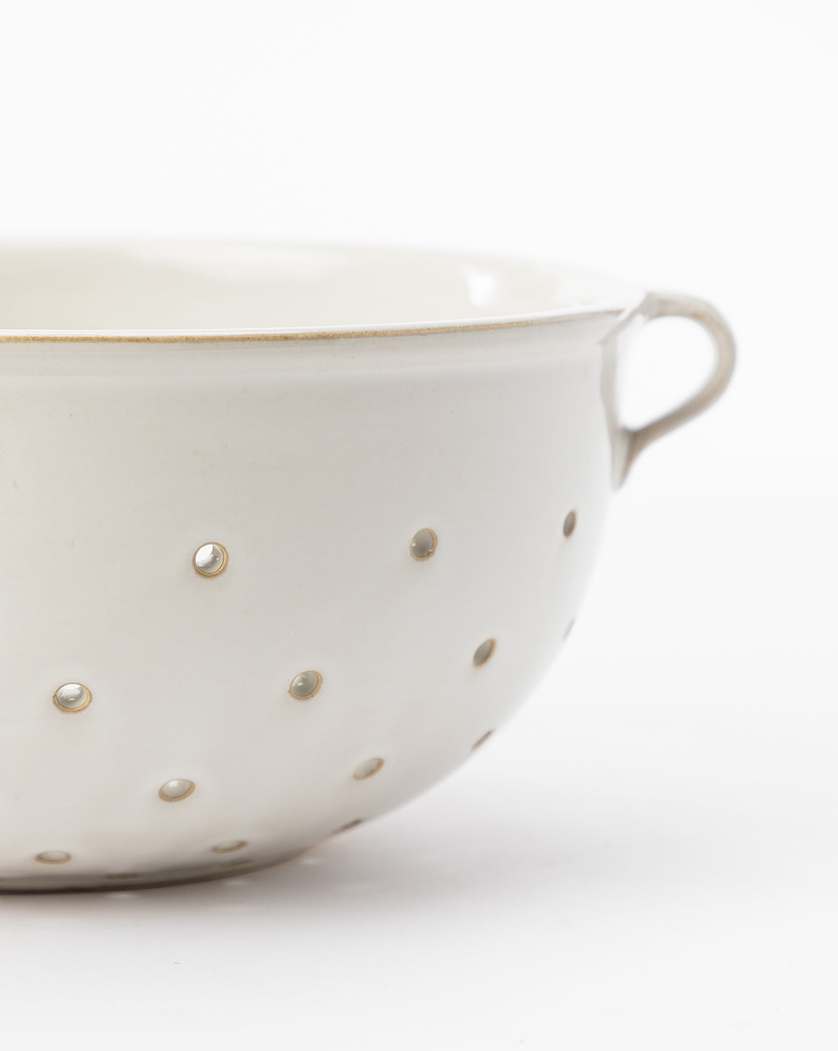 A close-up of the Glazed Berry Colander shows its white finish, small round holes, and single handle against a white background. This chic serveware elevates kitchen decor, with only part of the colander visible in the image, by McGee & Co.