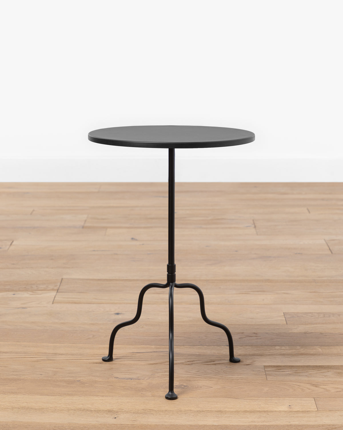 The Goodrich Iron Side Table, featuring a small round matte black top and slender three-legged base, stands on a light wood floor against a plain white background by McGee & Co.