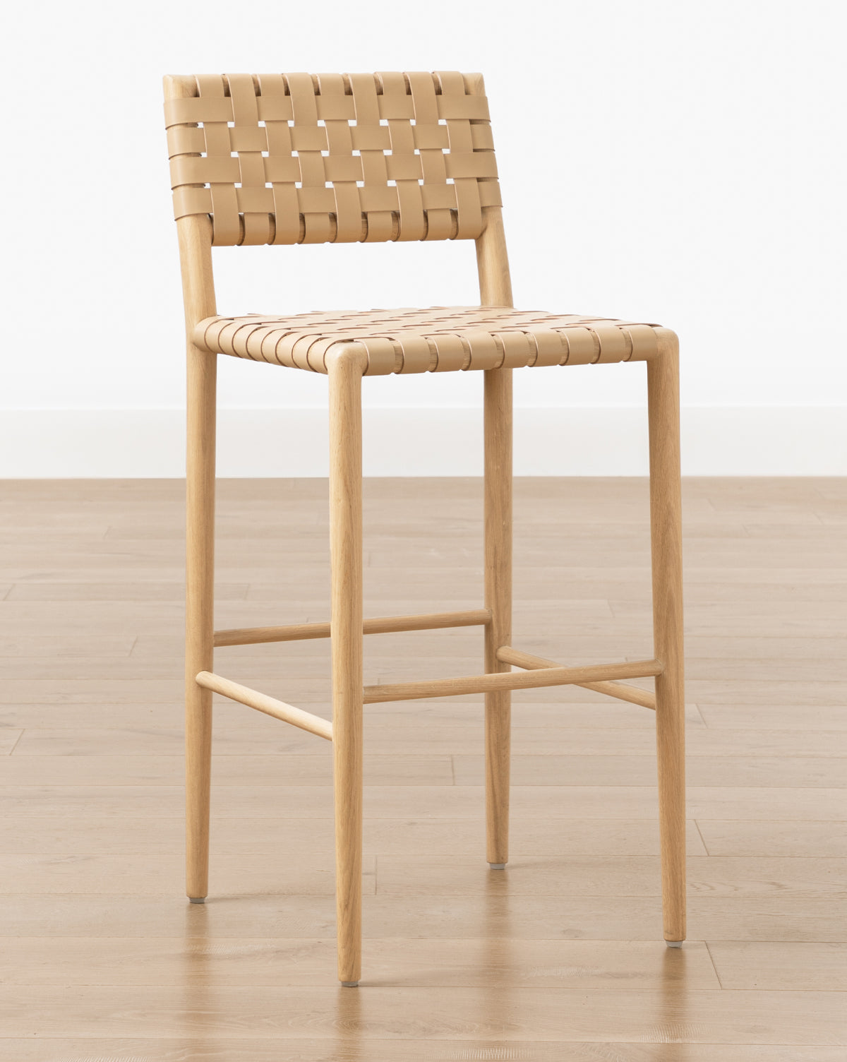 The Greely Stool features a white oak frame and a woven tan aniline-dyed leather seat and backrest, shown on a light wood floor against a plain white background by McGee & Co.