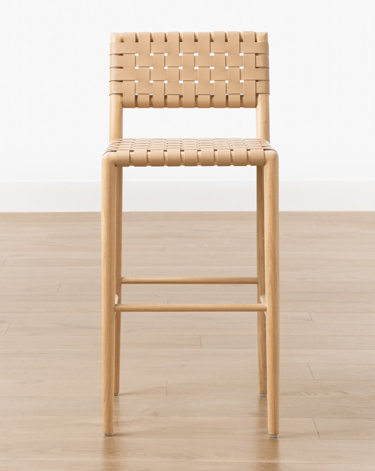 The Greely Stool features a light wood white oak frame with a woven aniline-dyed leather seat and backrest, pictured on a light wood floor against a plain white background by McGee & Co.