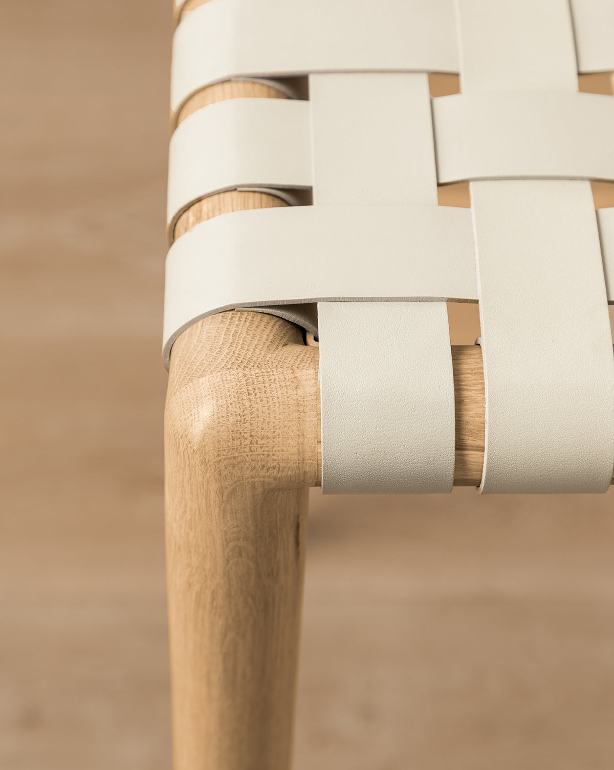 Close-up of the Greely Stools corner, showing its white oak frame and seat crafted from wide, aniline-dyed leather straps woven in a crisscross pattern. The blurred background emphasizes the stool’s detailed texture and craftsmanship by McGee & Co.