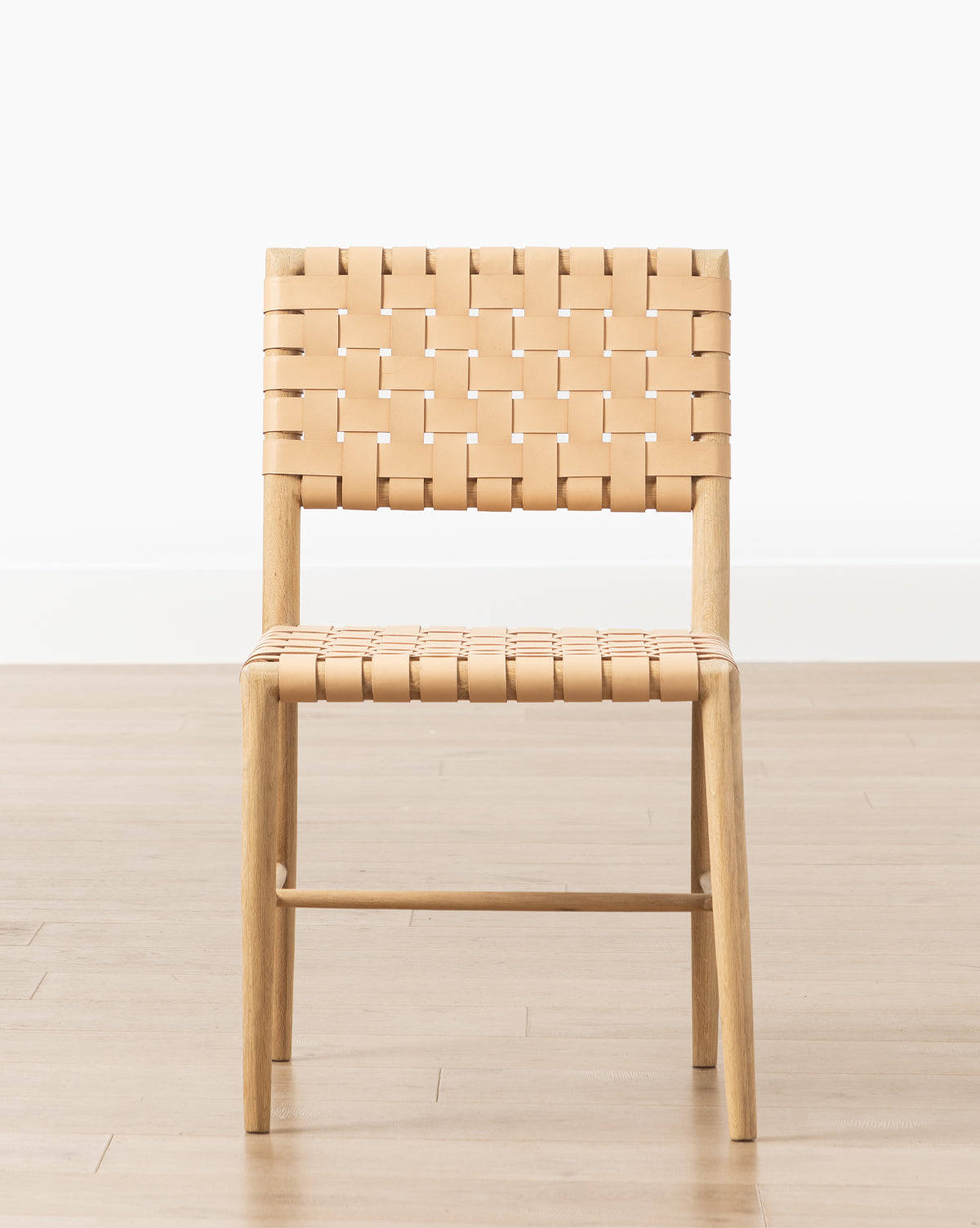 The Greely Dining Chair features a white oak frame with a light tan aniline-dyed leather woven seat and backrest, shown against a plain white wall and light wood floor, by McGee & Co.