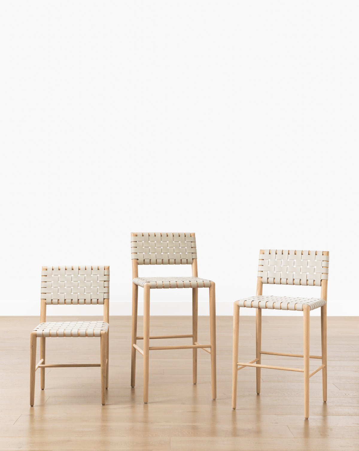 Three Greely Stools, each with a sturdy white oak frame and woven white seat and back, are arranged in a row on a wooden floor against a plain white background. The stools come in varying heights: short, medium, and tall, by McGee & Co.