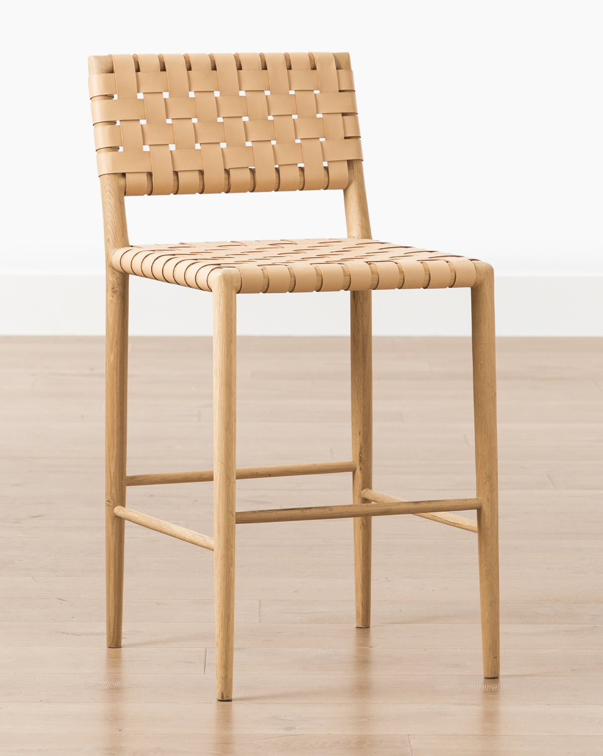 The Greely Stool features a woven, light tan aniline-dyed leather seat and backrest atop wood, shown on a light wooden floor against a plain white background by McGee & Co.