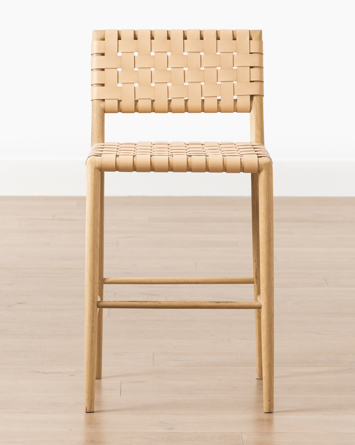 The Greely Stool features a white oak frame with a woven tan aniline-dyed leather seat and backrest, shown on a light wooden floor against a plain white wall, by McGee & Co.
