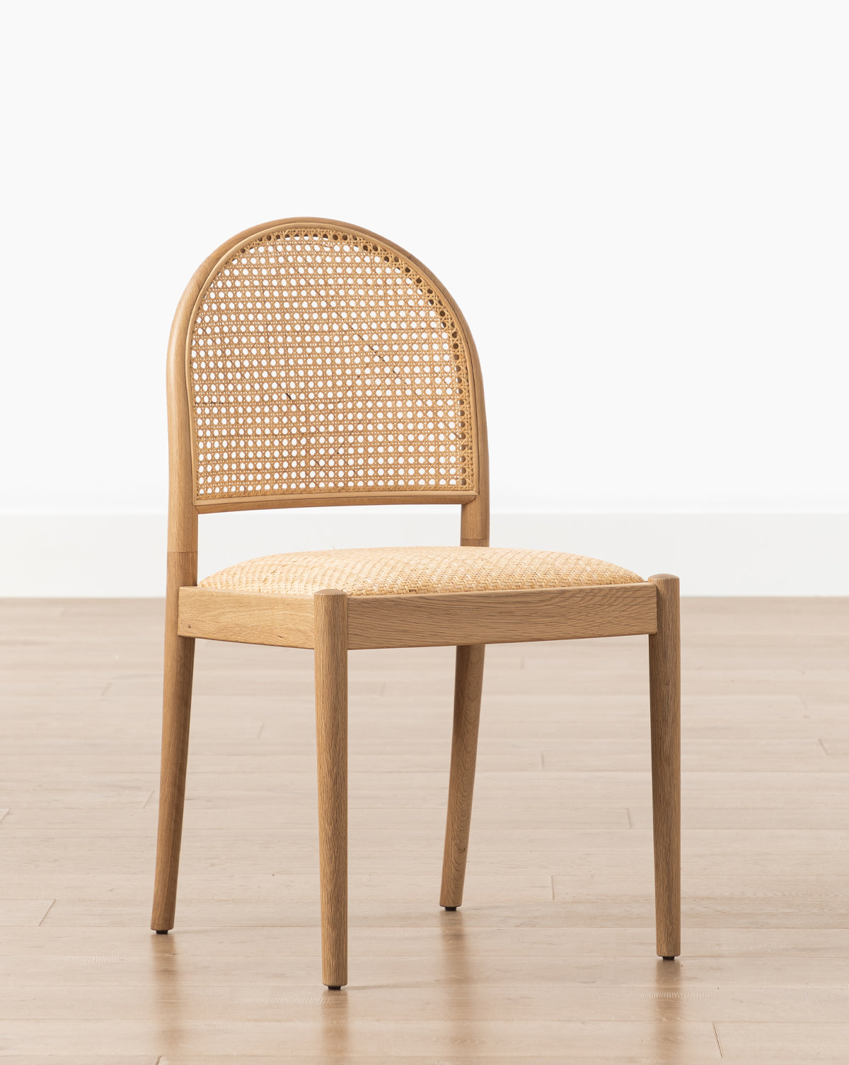 The Hadden Natural Dining Chair features a curved, woven cane back and matching seat in natural oak, shown on a light wood floor with a plain white wall background by McGee & Co.