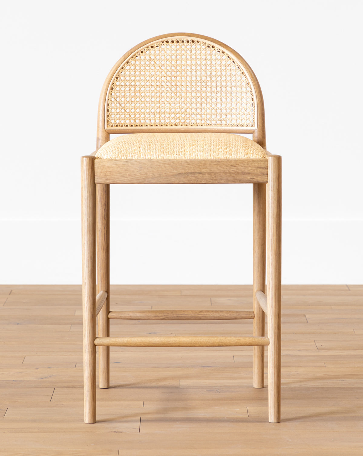 The Hadden Natural Counter Stool features an oak frame, woven rattan seat and backrest, rounded back, and straight legs—ideal for coastal décor—shown here on a light wooden floor against a plain white wall by McGee & Co.