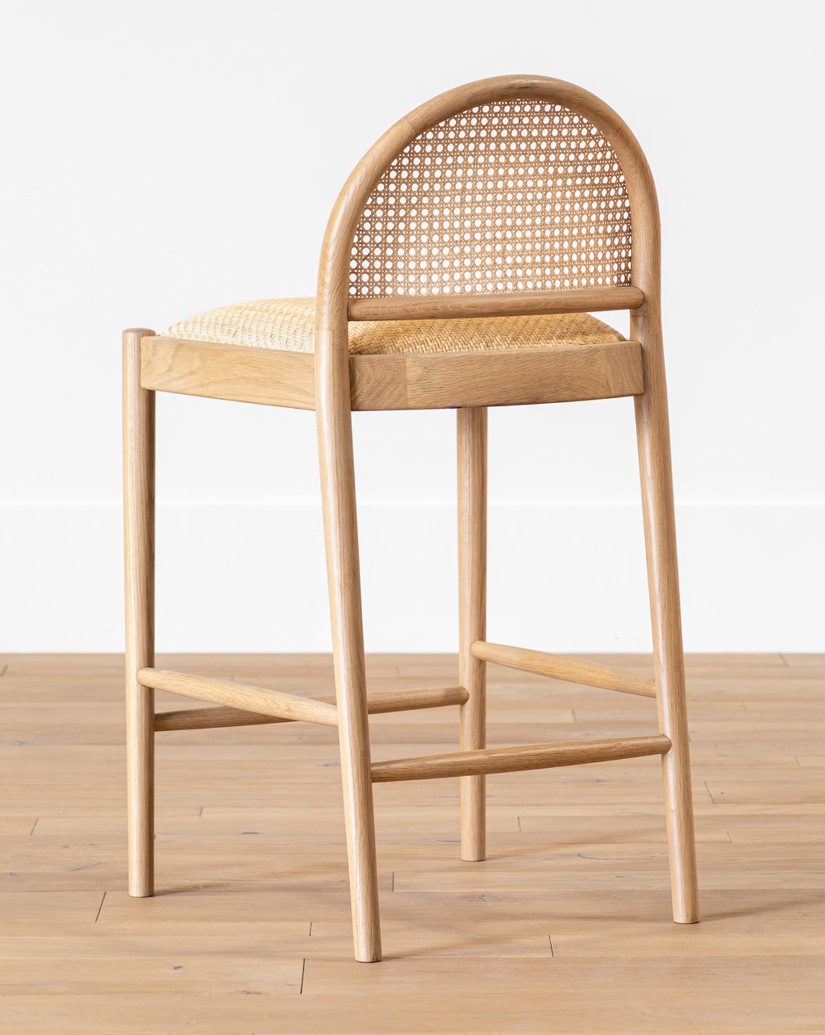 The Hadden Natural Counter Stool, with an oak frame and woven cane backrest and seat, features rounded edges and four straight legs. Set on a light wooden floor against a plain white wall, its ideal for coastal-inspired interiors by McGee & Co.