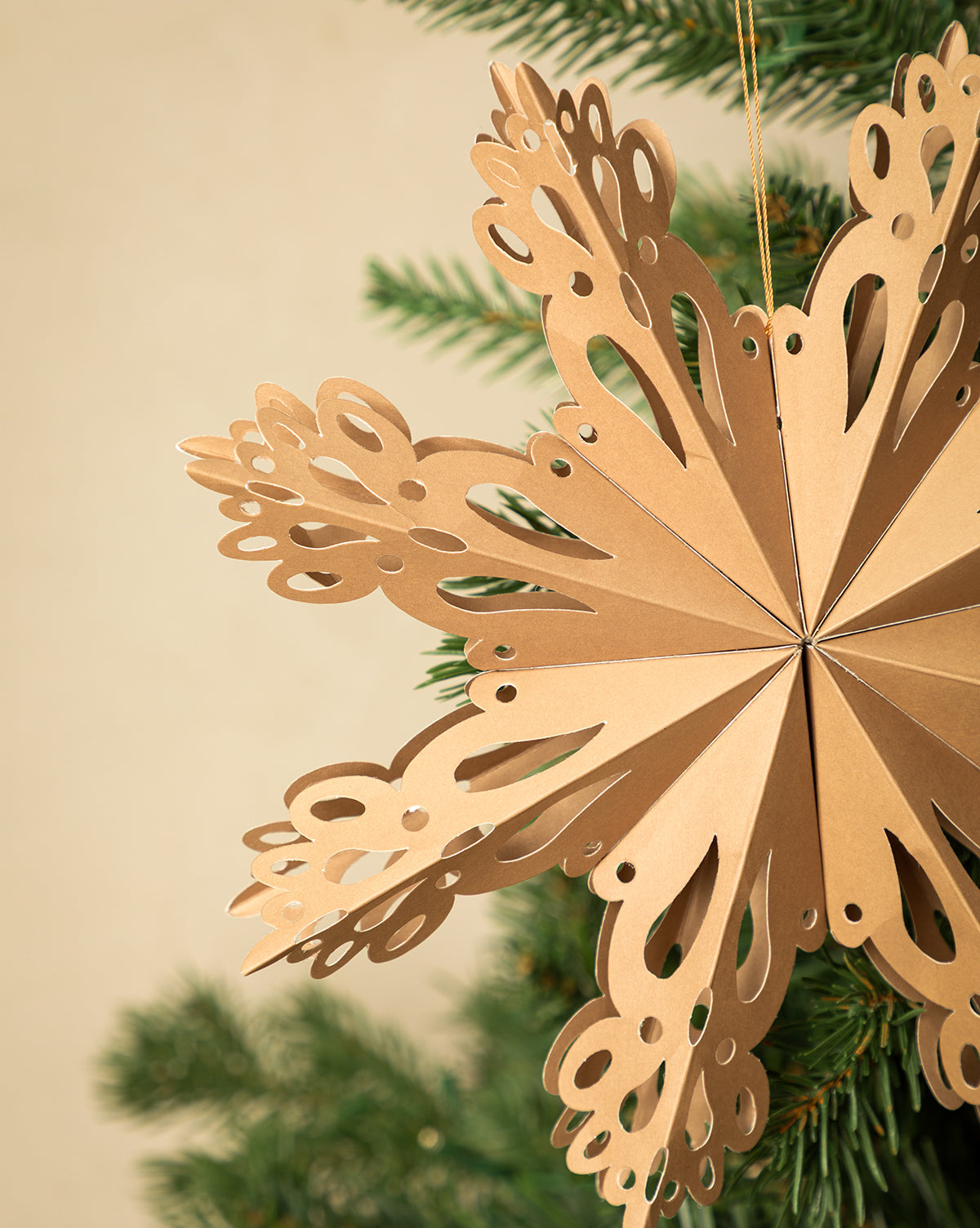 A set of three Handmade Assorted Recycled Paper Snowflakes adds eco-friendly charm and neutral hues to your holiday decor, hanging beautifully on a green Christmas tree branch against a simple beige background, by McGee & Co.