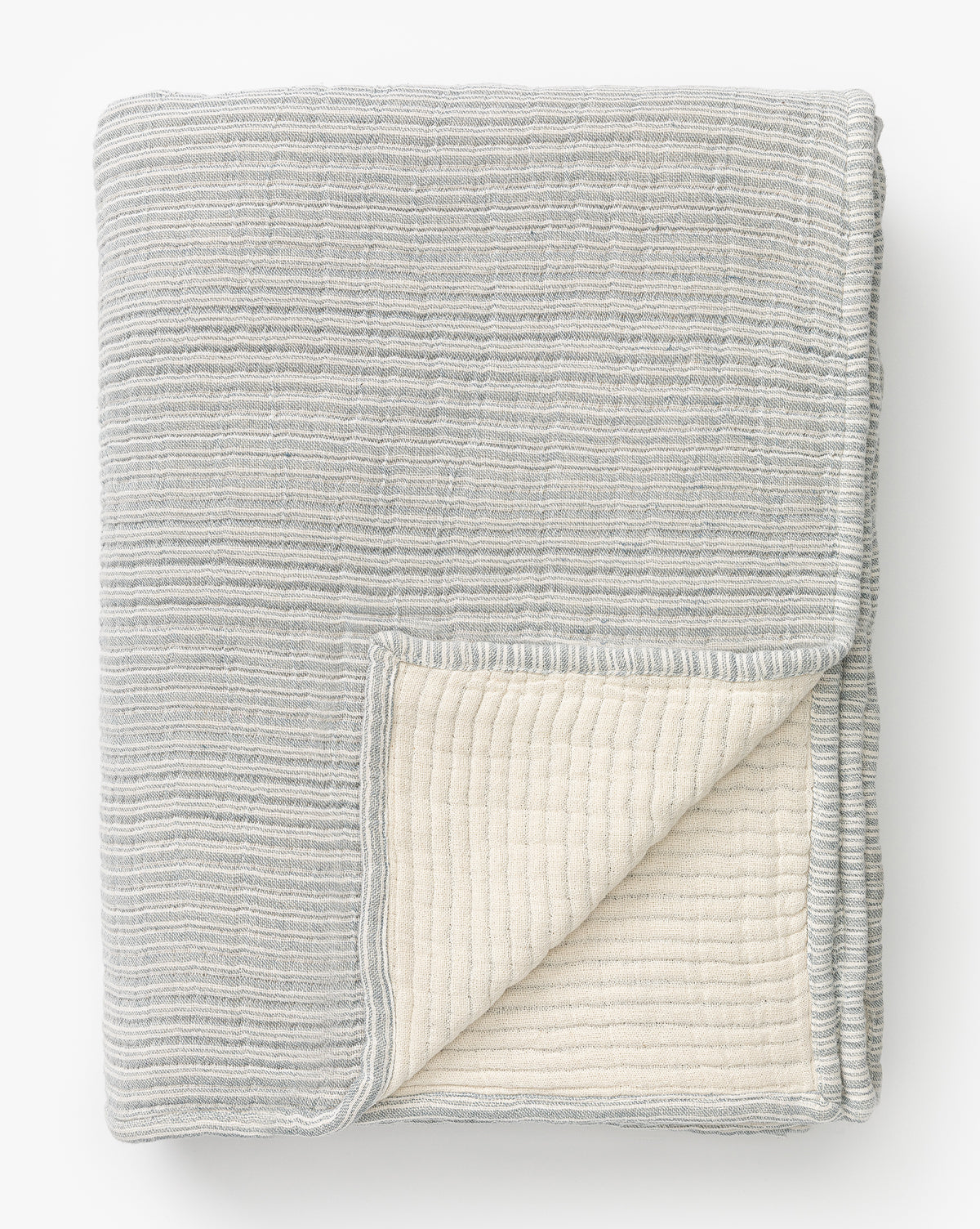 The Hanson Striped Cotton Coverlet features thin horizontal stripes in light gray and cream, with a textured design. One corner is flipped to show the contrasting cream underside, giving this neutral bedding a soft and cozy look.