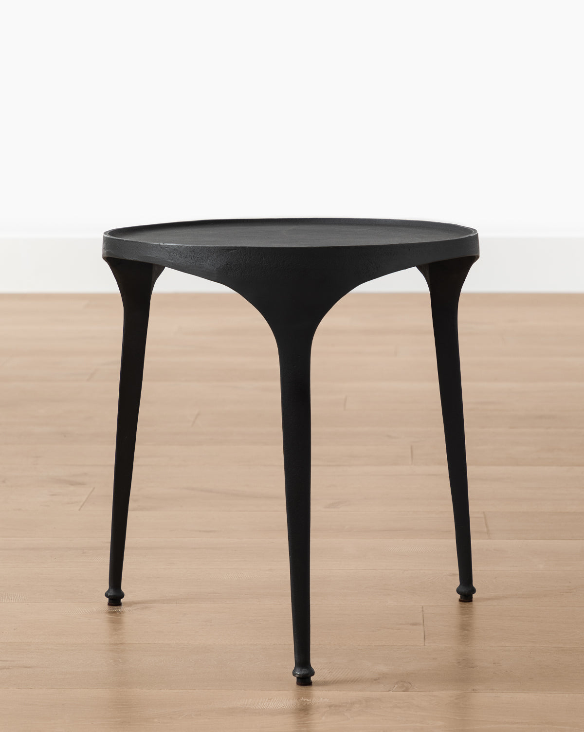 The Haskell Side Table, small and round with three slender legs, stands on a light wood floor by a plain white wall, highlighting its sleek contemporary design, by McGee & Co.