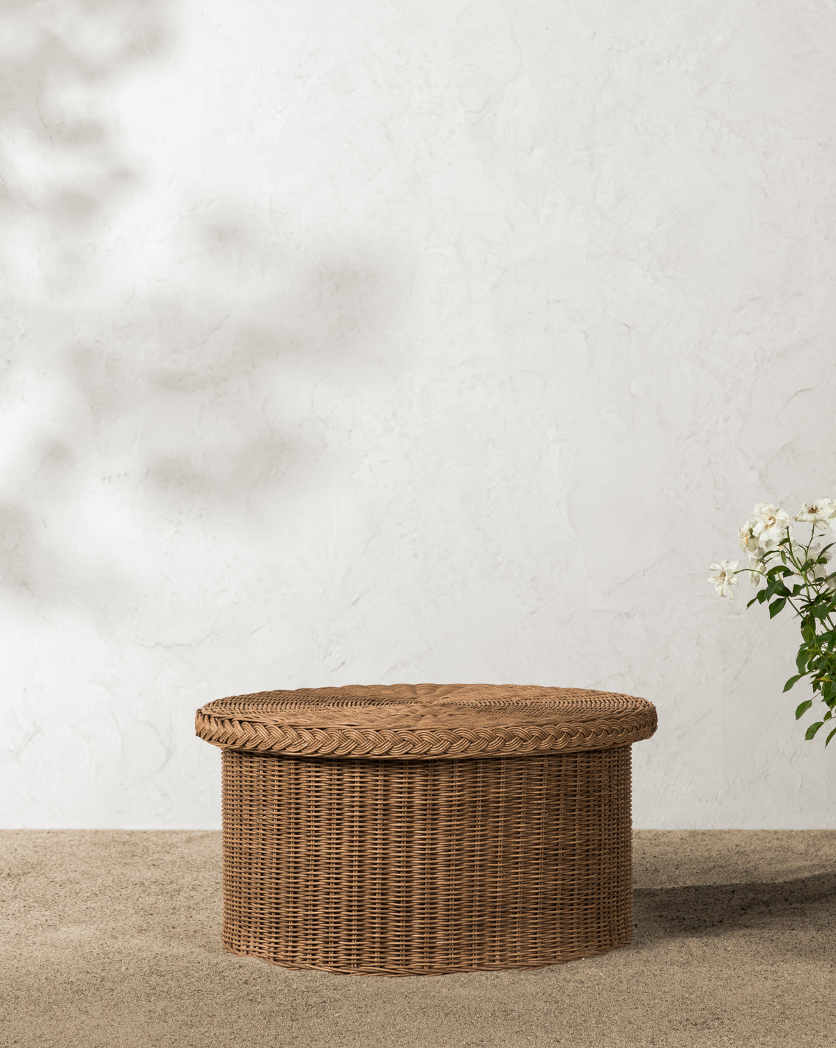 The Haviland Coffee Table, featuring a weather-resistant wicker woven design, sits on small pebbles by a textured white wall as soft natural light casts gentle shadows by McGee & Co.