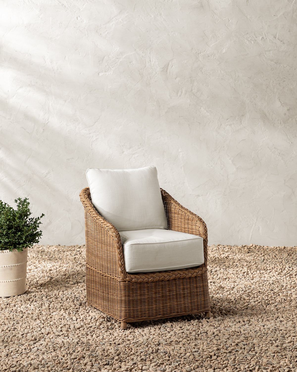 The Haviland Outdoor Dining Chair with weather-resistant wicker and white cushions is placed on a textured pebble floor beside a potted plant against a light, textured wall by McGee & Co.
