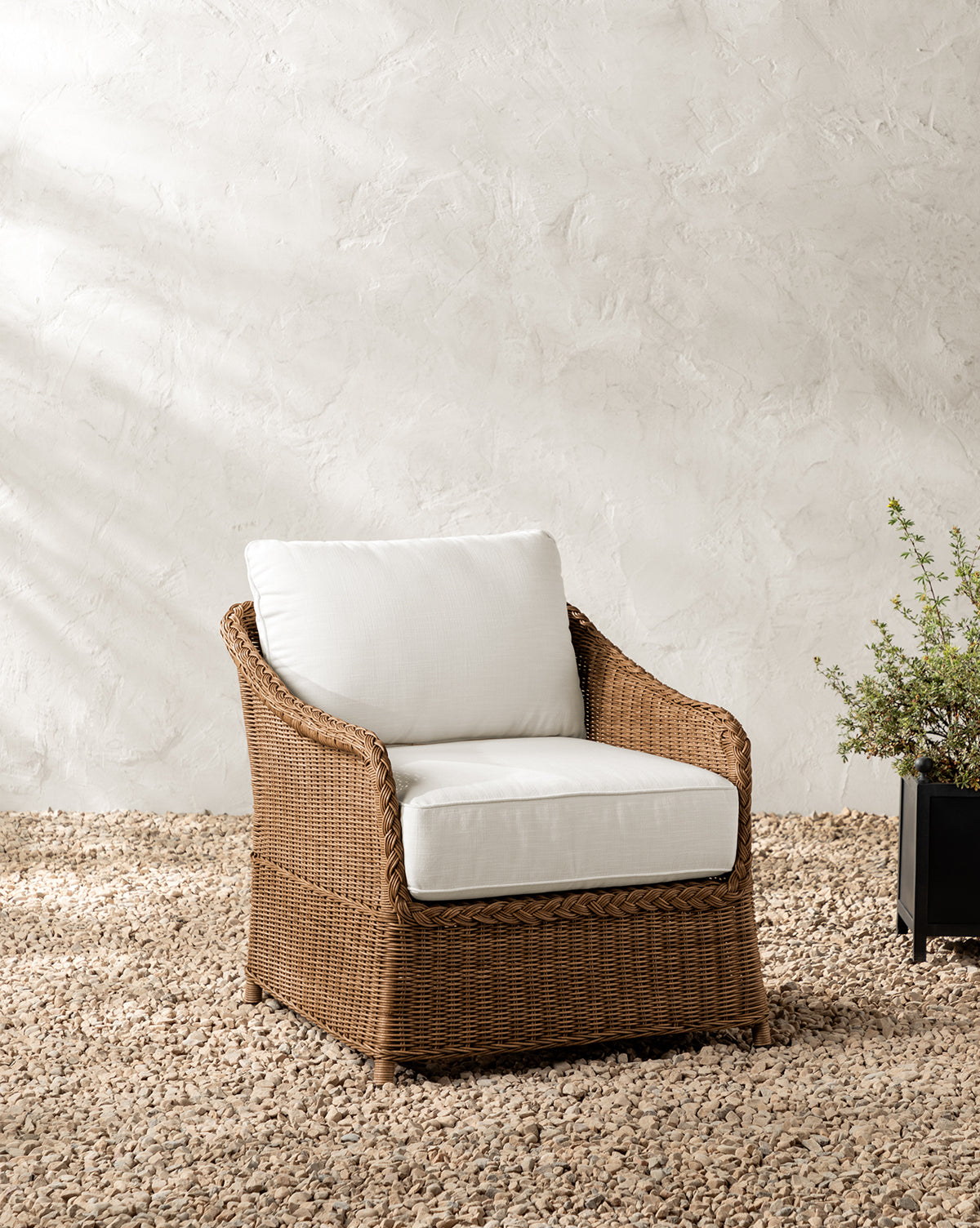 The Haviland Outdoor Lounge Chair with weather-resistant wicker and white cushions sits on gravel near a black planter with greenery, set against a textured off-white wall, by McGee & Co.