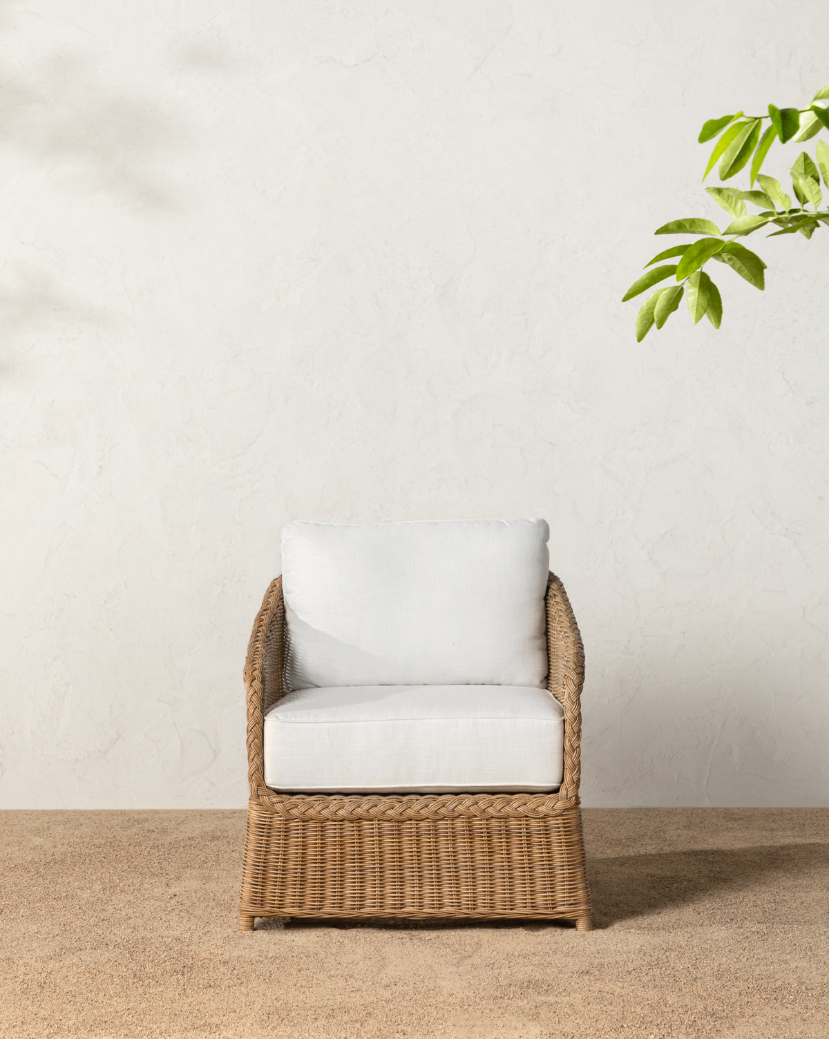 The Haviland Outdoor Lounge Chair, featuring weather-resistant wicker and white cushions, is placed on small pebbles before a textured white wall, beside a black planter filled with lush green foliage, by McGee & Co.