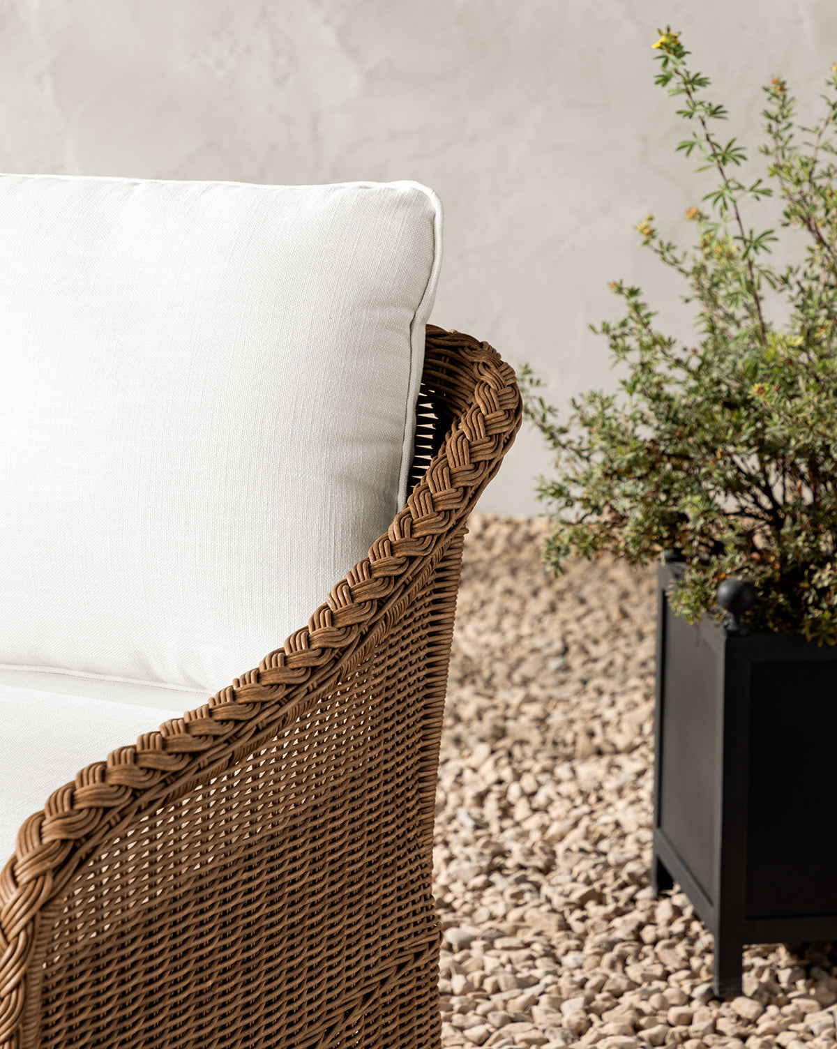 The Haviland Outdoor Lounge Chair, crafted from weather-resistant wicker with white cushions, sits on gravel beside a potted plant in a black rectangular planter. A neutral textured wall provides the backdrop, by McGee & Co.