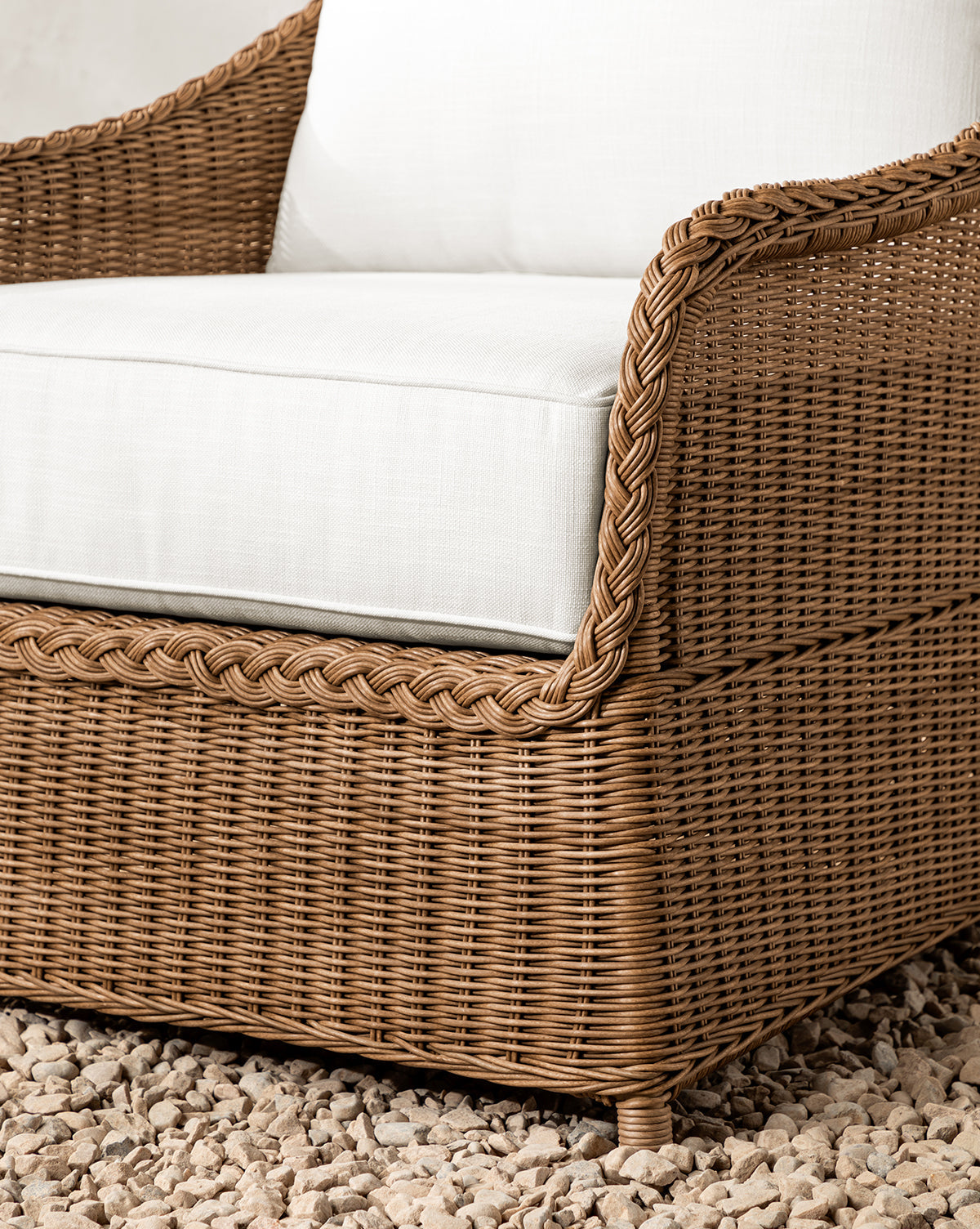 Close-up of the Haviland Outdoor Lounge Chair, made from brown weather-resistant wicker with braided details and white cushioned seat and backrest, placed on a bed of small, light-colored stones by McGee & Co.
