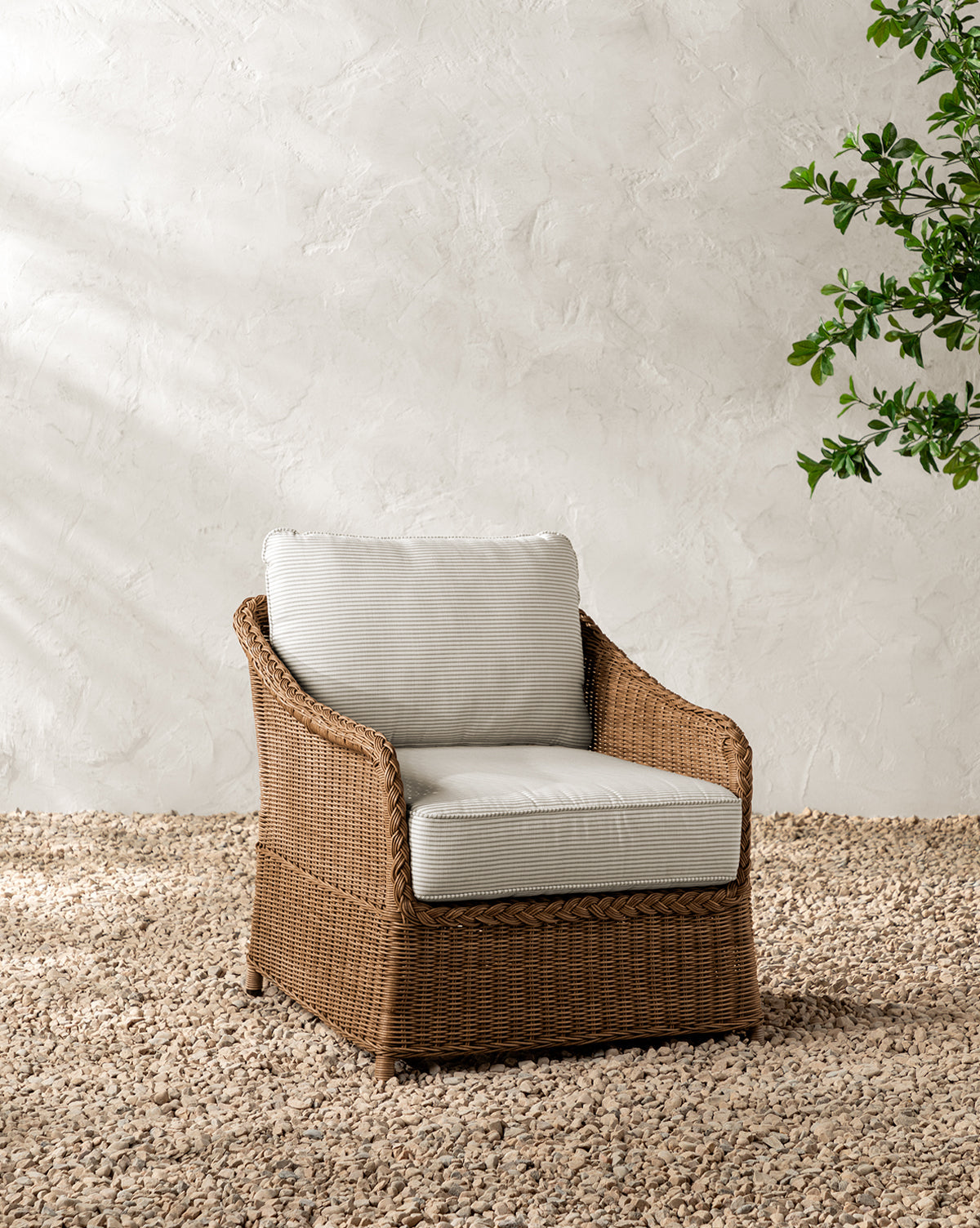 The Haviland Outdoor Lounge Chair with Perennials Taupe Stripe sits on small pebbles against a textured white wall, with green leafy branches visible in the top right corner, by McGee & Co.