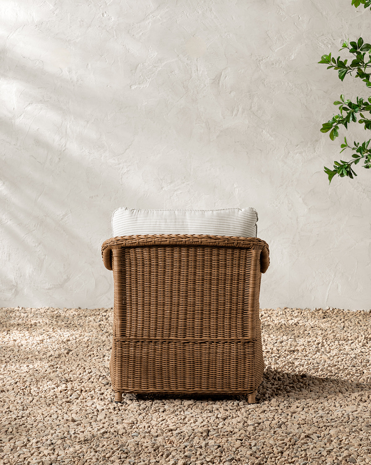 The Haviland Outdoor Lounge Chair with Perennials Taupe Stripe, featuring a weather-resistant wicker frame and light cushion, sits on gravel by a textured white wall with sunlight and a green leafy branch to the right by McGee & Co.