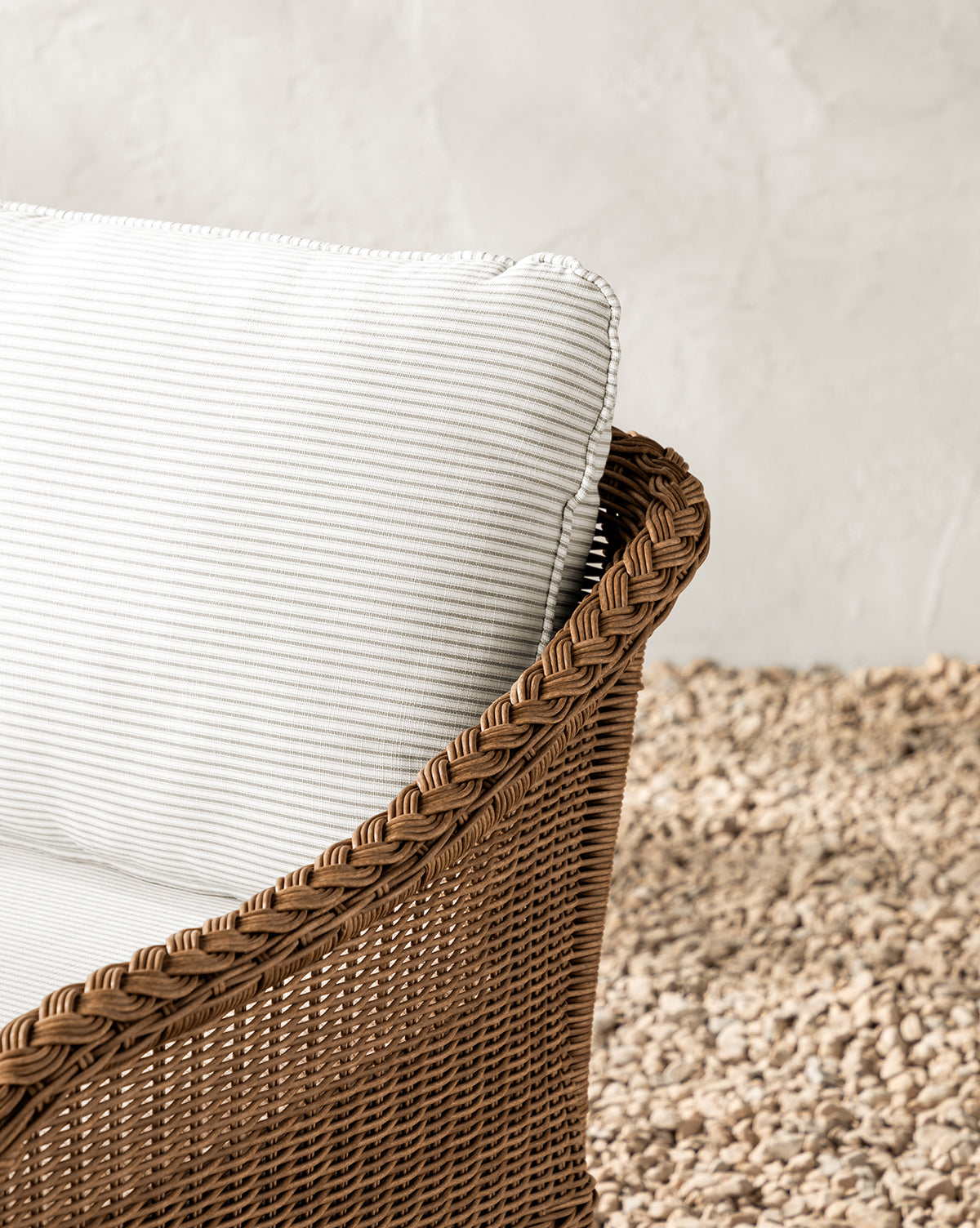 Close-up of the Haviland Outdoor Lounge Chair with Perennials Taupe Stripe cushion, featuring weather-resistant wicker, set on a textured pebble rug against a neutral, textured wall backdrop, by McGee & Co.
