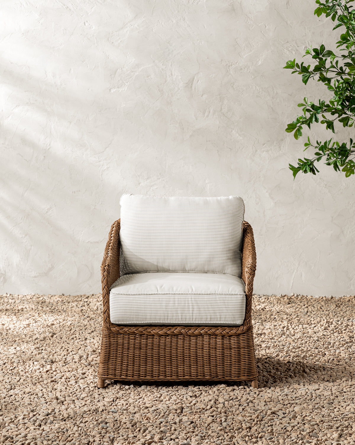 The Haviland Outdoor Lounge Chair with Perennials Taupe Stripe sits on a gravel surface before a textured light beige wall, with leafy green branches visible in the upper right corner, by McGee & Co.