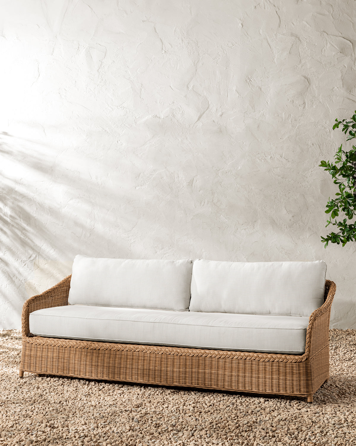 The Haviland Outdoor Sofa with Perennials Taupe Stripe 88 features weather-resistant white cushions and sits on a pebble floor near a textured white wall, with a leafy plant visible to the right, by McGee & Co.