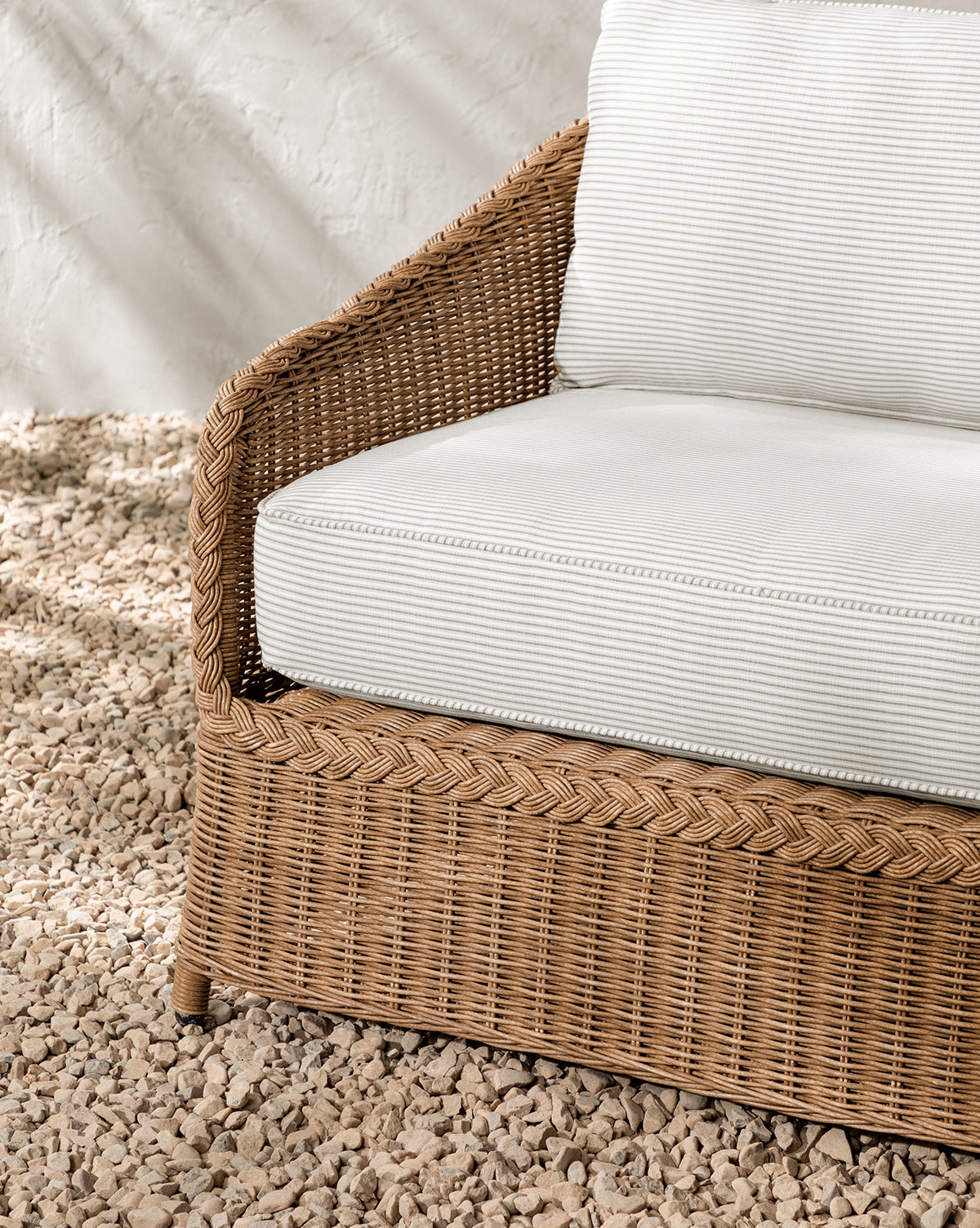 A close-up of the Haviland Outdoor Sofa with Perennials Taupe Stripe 88, featuring cream-colored cushions, is set on light pebbles against a textured off-white wall, by McGee & Co.