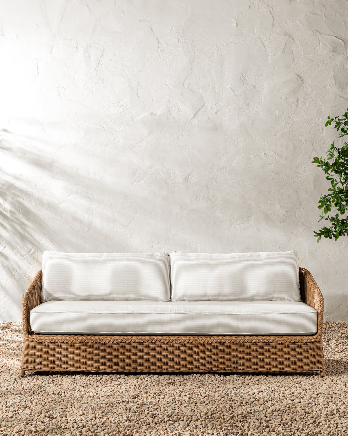 A Haviland Outdoor Sofa with Perennials Taupe Stripe 88 sits on a textured rug against a white, stucco-textured wall, with a green plant to the right and soft sunlight casting gentle shadows from the left by McGee & Co.