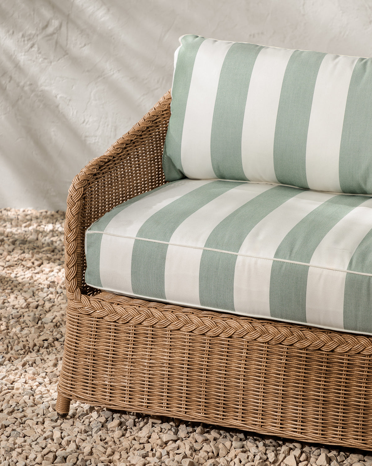 The Haviland Outdoor Sofa with Striped Cushions 88 rests on small white pebbles against a textured white wall, while sunlight casts soft shadows over its green and white cushions, by McGee & Co.