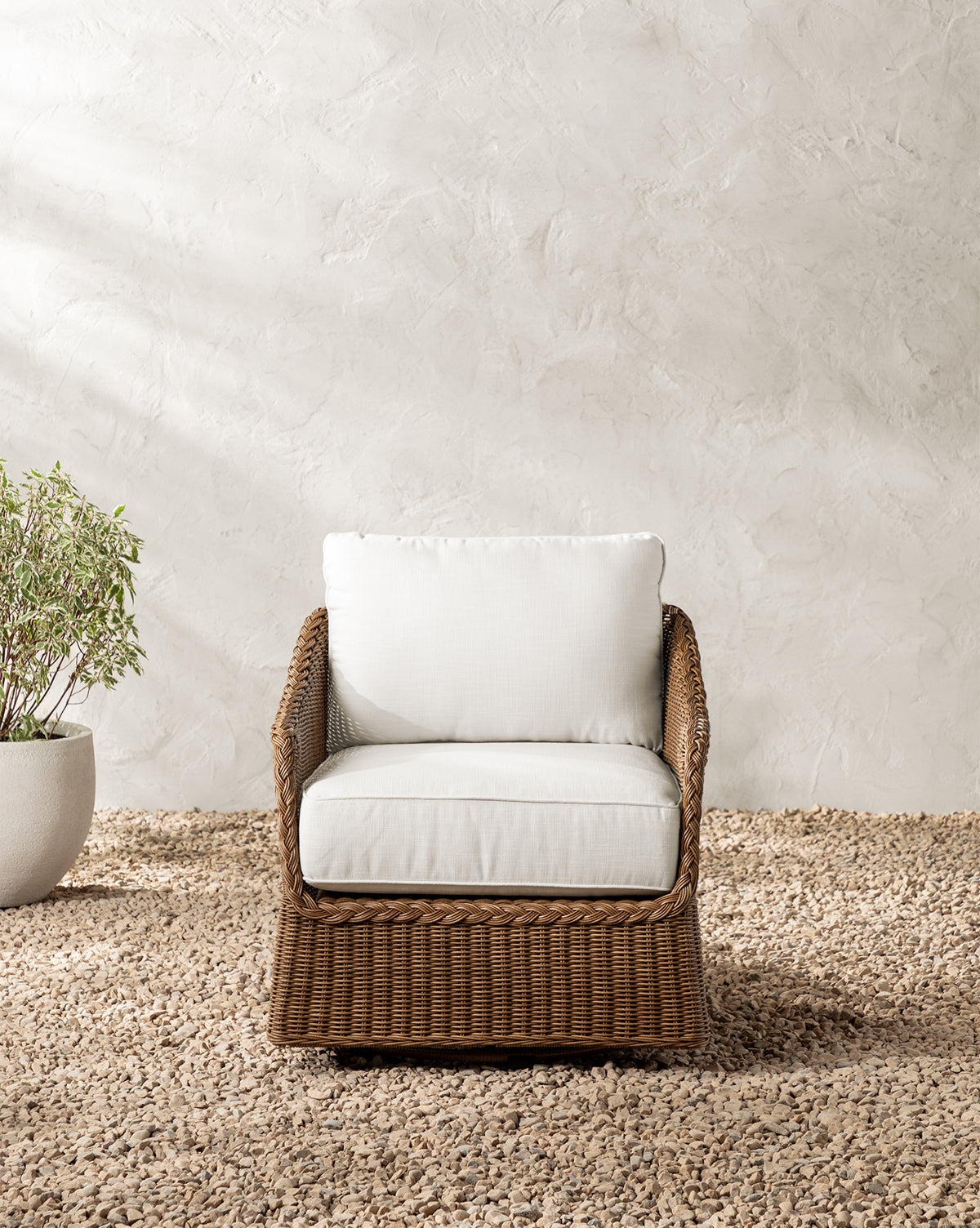 The Haviland Outdoor Swivel Lounge Chair with white cushions sits on gravel by a potted plant, set against a textured light beige wall, by McGee & Co.