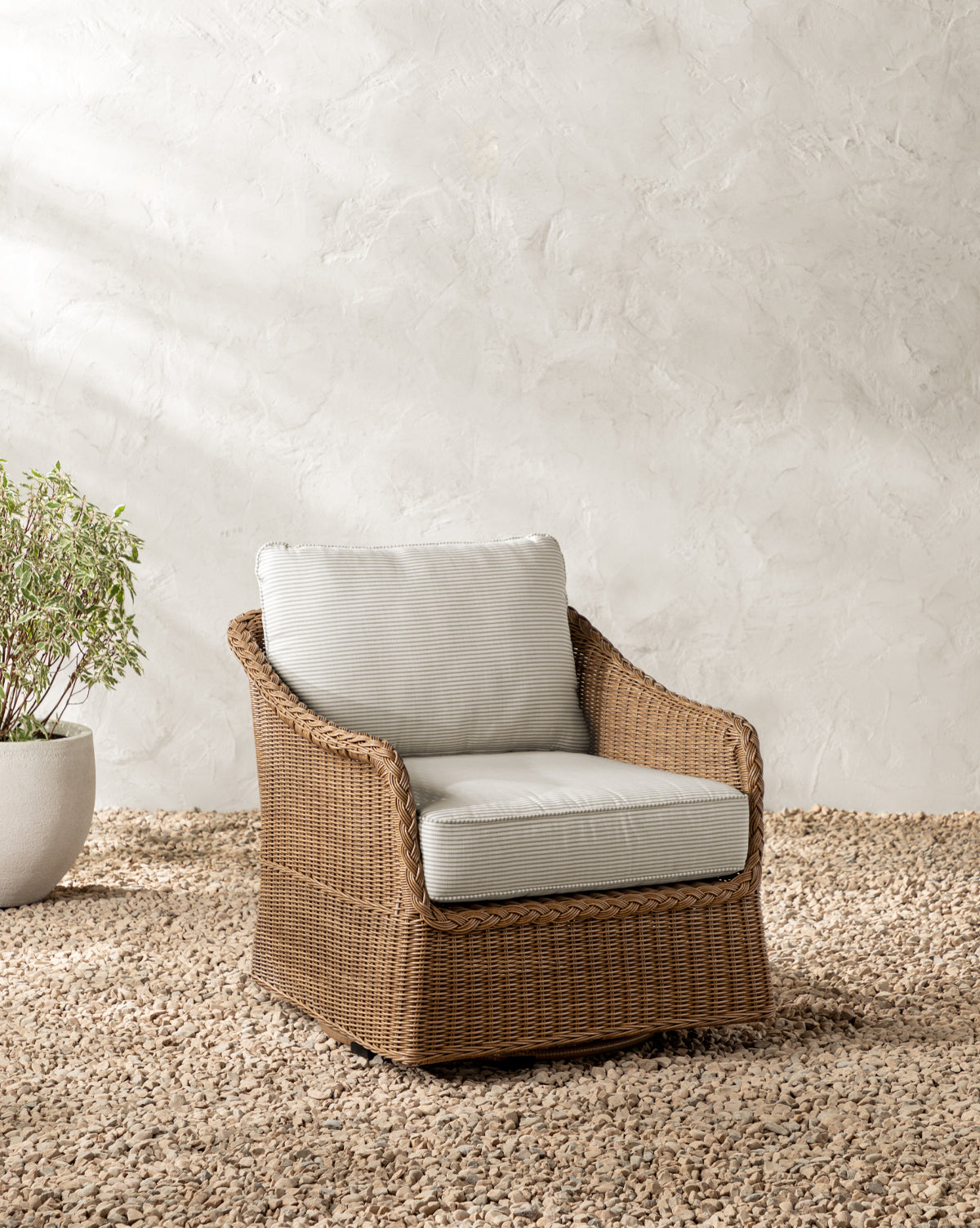 The Haviland Outdoor Swivel Lounge Chair with light gray cushions rests on a pebble floor beside a potted plant, set against a textured off-white wall, by McGee & Co.