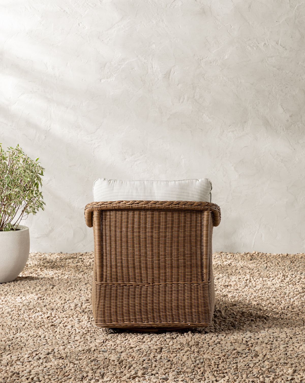 The Haviland Outdoor Swivel Lounge Chair with a light cushion sits on a pebble-covered ground near a textured white wall, with a potted green plant partially visible on the left, by McGee & Co.