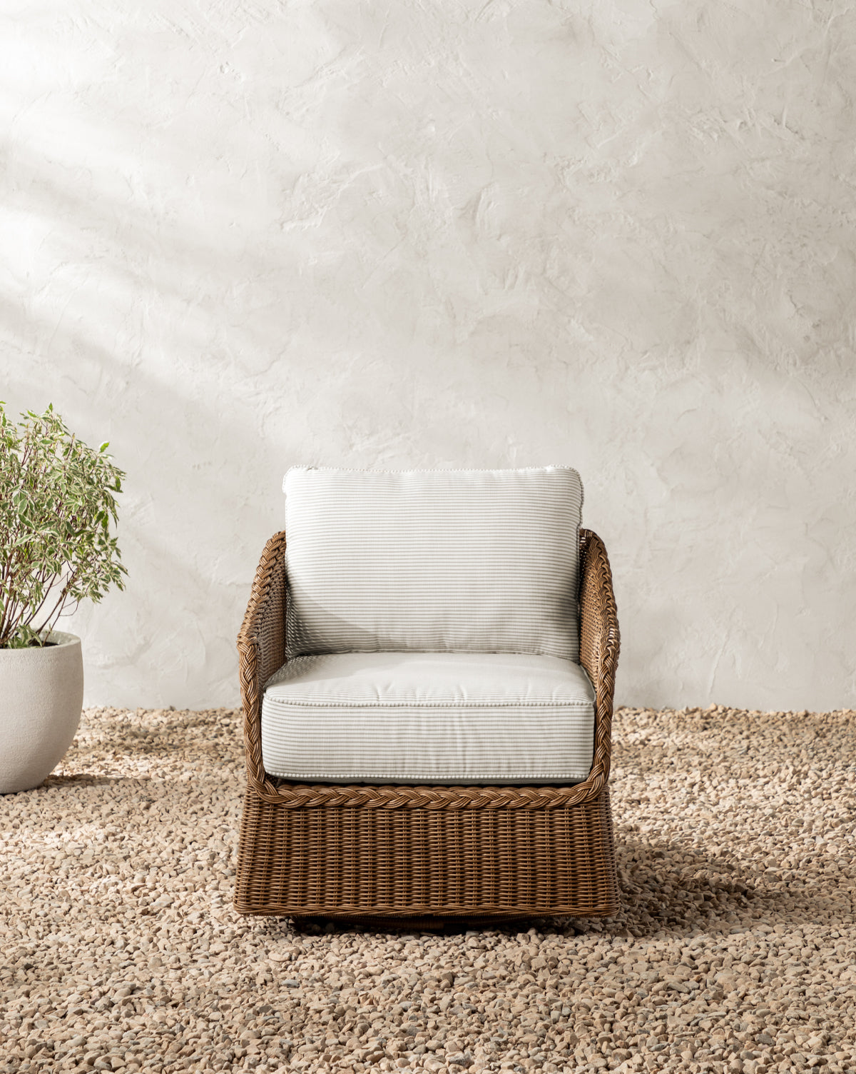 The Haviland Outdoor Swivel Lounge Chair with white cushions is placed on a pebble-covered ground beside a potted plant, set against a textured light-colored wall by McGee & Co.