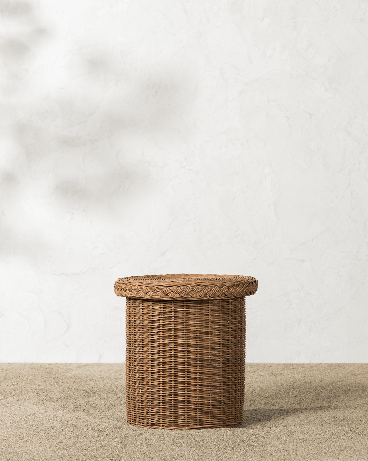 The Haviland Side Table, a round wicker basket with a lid, stands on gravel by a textured white wall. Its weather-resistant design and warm sunlight make it perfect for outdoor lounging and versatile as both storage and side table, by McGee & Co.