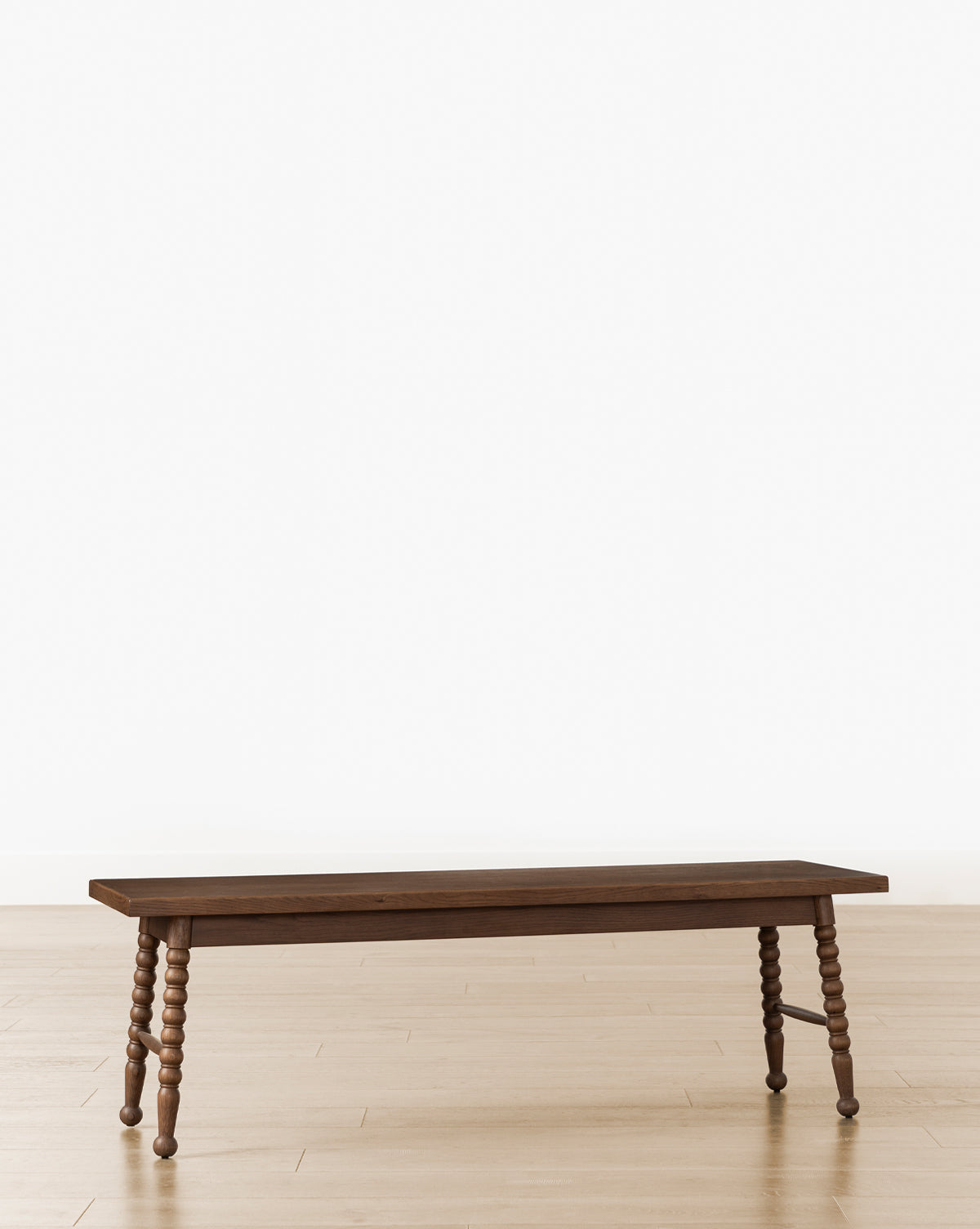 The Heathcliff Bench features a smooth wooden top and four intricately carved legs, shown on a light wood floor with a plain white background, by McGee & Co.