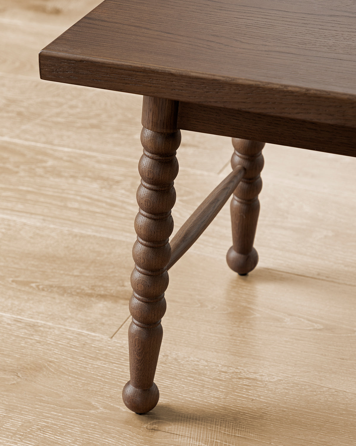 Close-up of the Heathcliff Benchs wooden, turned decorative leg connected to a rectangular tabletop, positioned on a light wood floor, by McGee & Co.