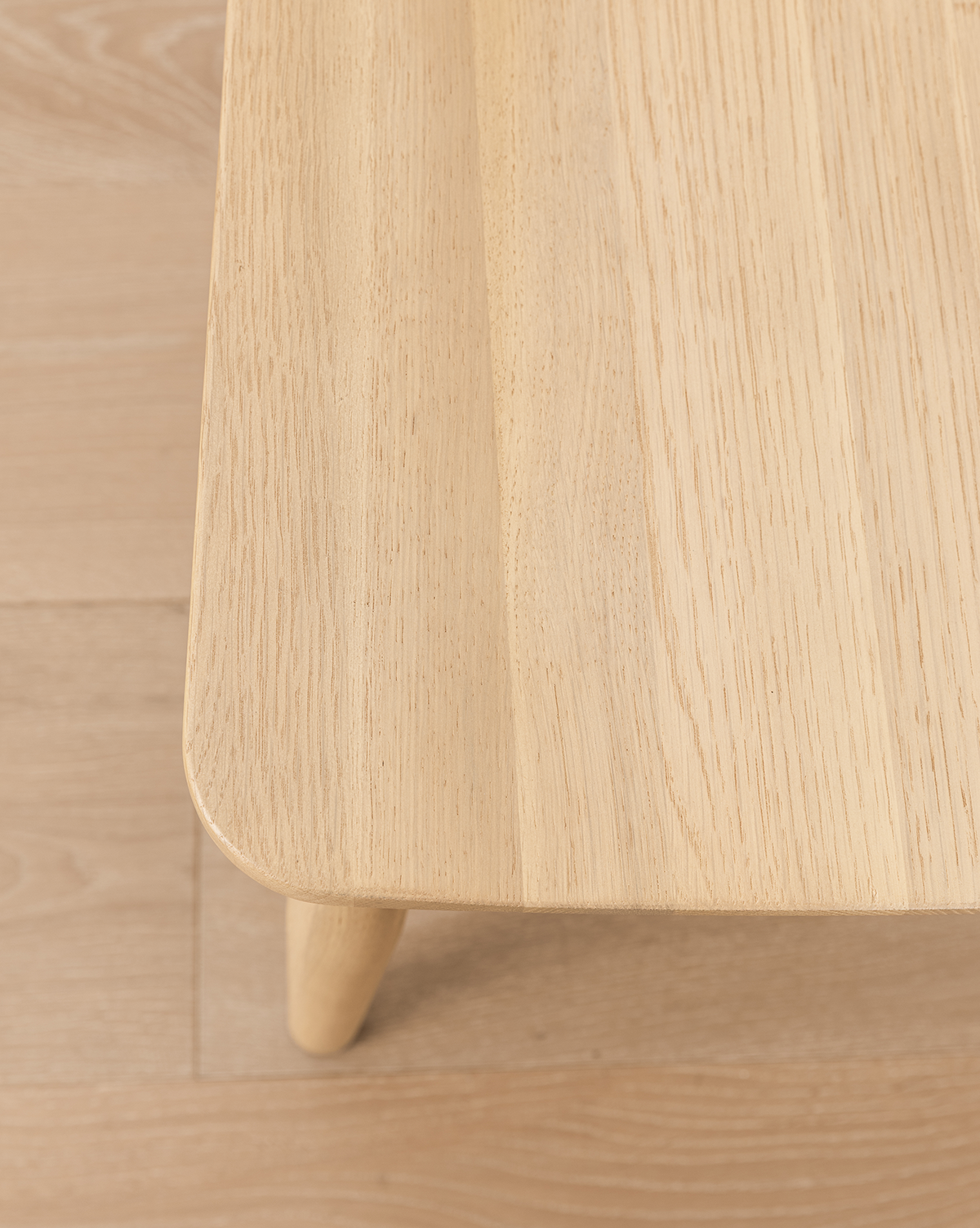 Close-up of a light wooden table with rounded corners and visible grain, set on a matching floor next to a versatile Hemingway Chair, by McGee & Co.