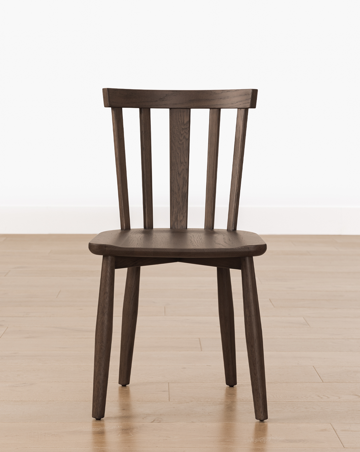 A Hemingway Chair with a dark wood finish, slatted backrest, and angled legs sits on a light wood floor against a plain white wall, by McGee & Co.