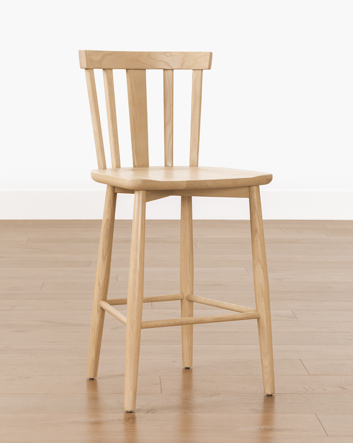A simple solid wood Hemingway Stool with a slatted backrest and four angled legs, set on a light wood floor against a plain white wall—ideal for pairing with kitchen counters or as an alternative to classic seating options, by McGee & Co.