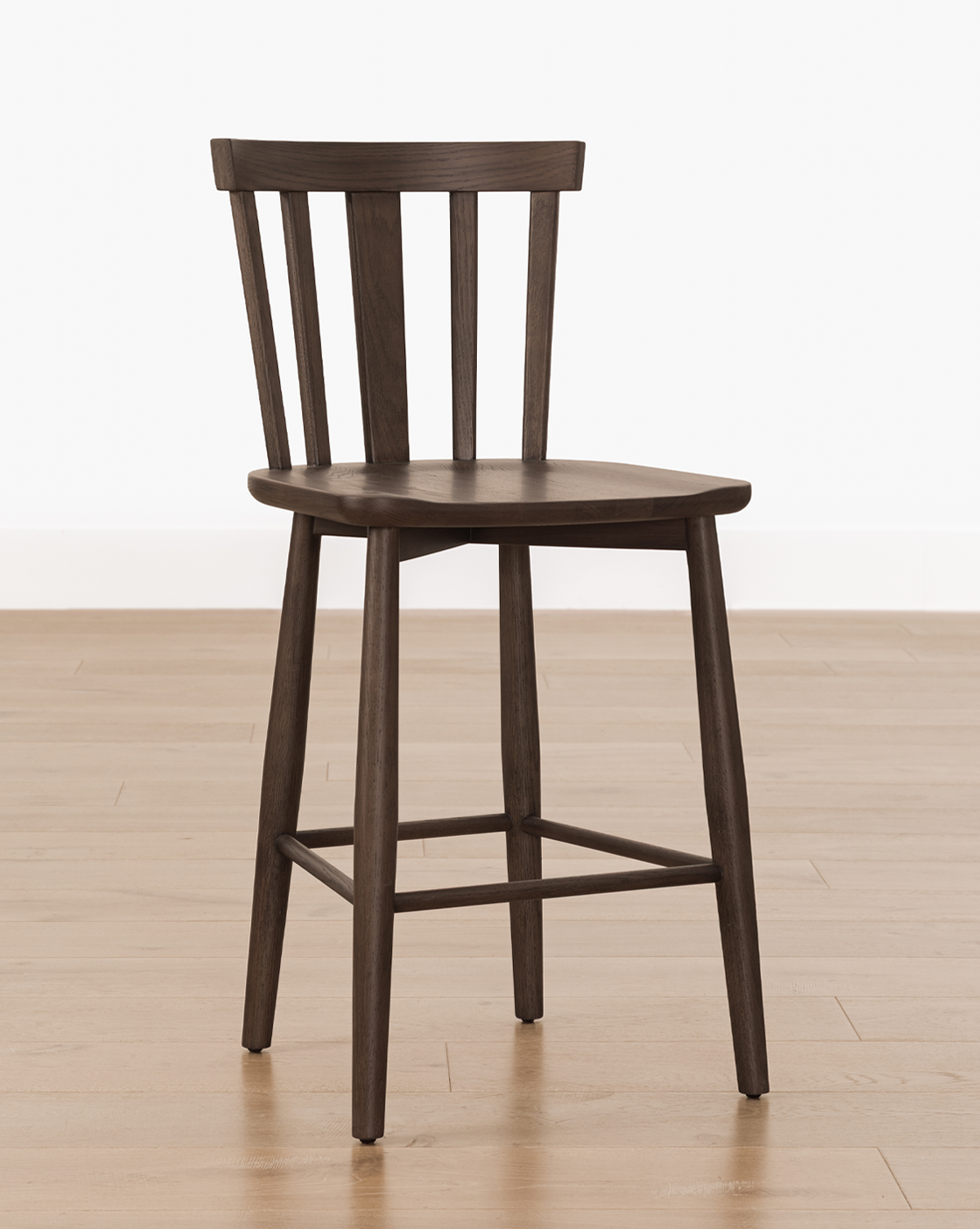 The Hemingway Stool, a dark solid wood stool with a flat seat, slightly angled legs, and a backrest featuring three vertical slats, sits on a light wood floor against a plain white wall—ideal for pairing with kitchen counters, by McGee & Co.