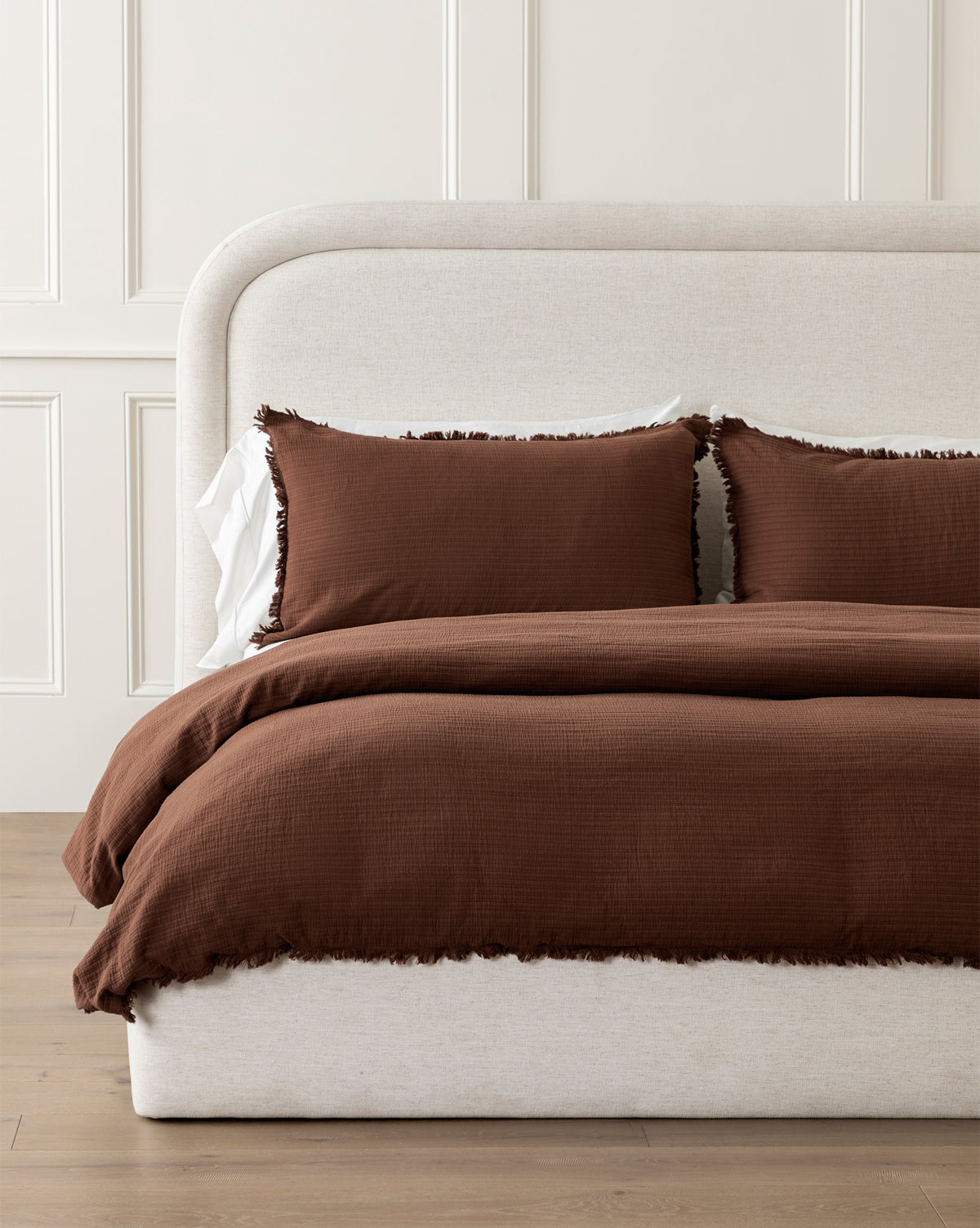 A bed with a light beige upholstered headboard and frame is dressed in the Hendricks Chocolate Cotton Duvet Cover and matching shams, set in a room with white paneled walls and wooden flooring.