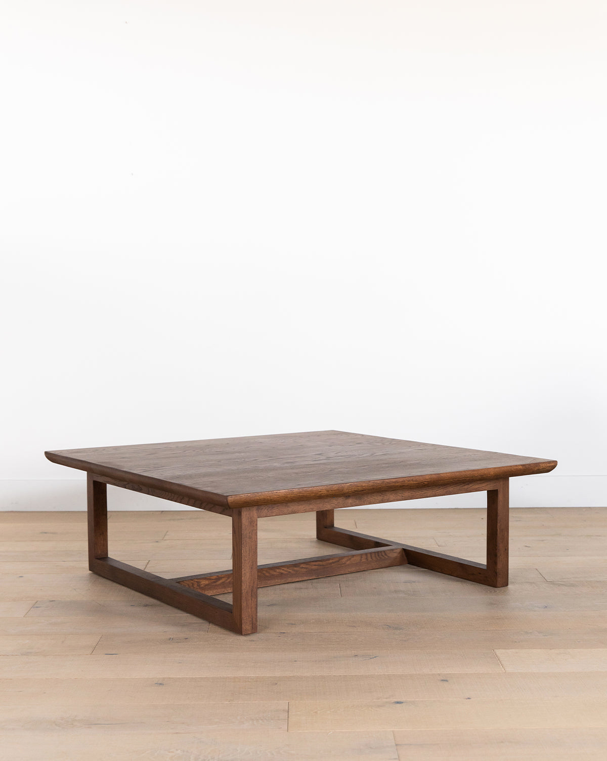 The Herrin Coffee Table is a modern, square coffee table made from solid oak, with a flat top and open rectangular legs, shown on a light wood floor in front of a plain white wall, by McGee & Co.