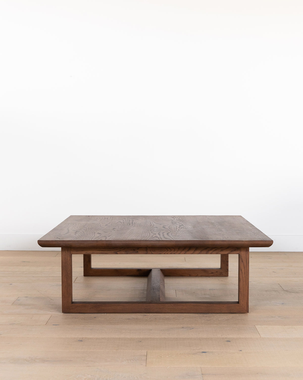 The Herrin Coffee Table is a modern solid oak piece with a rectangular top and geometric base, showcased on a light wood floor beside a plain white wall, by McGee & Co.