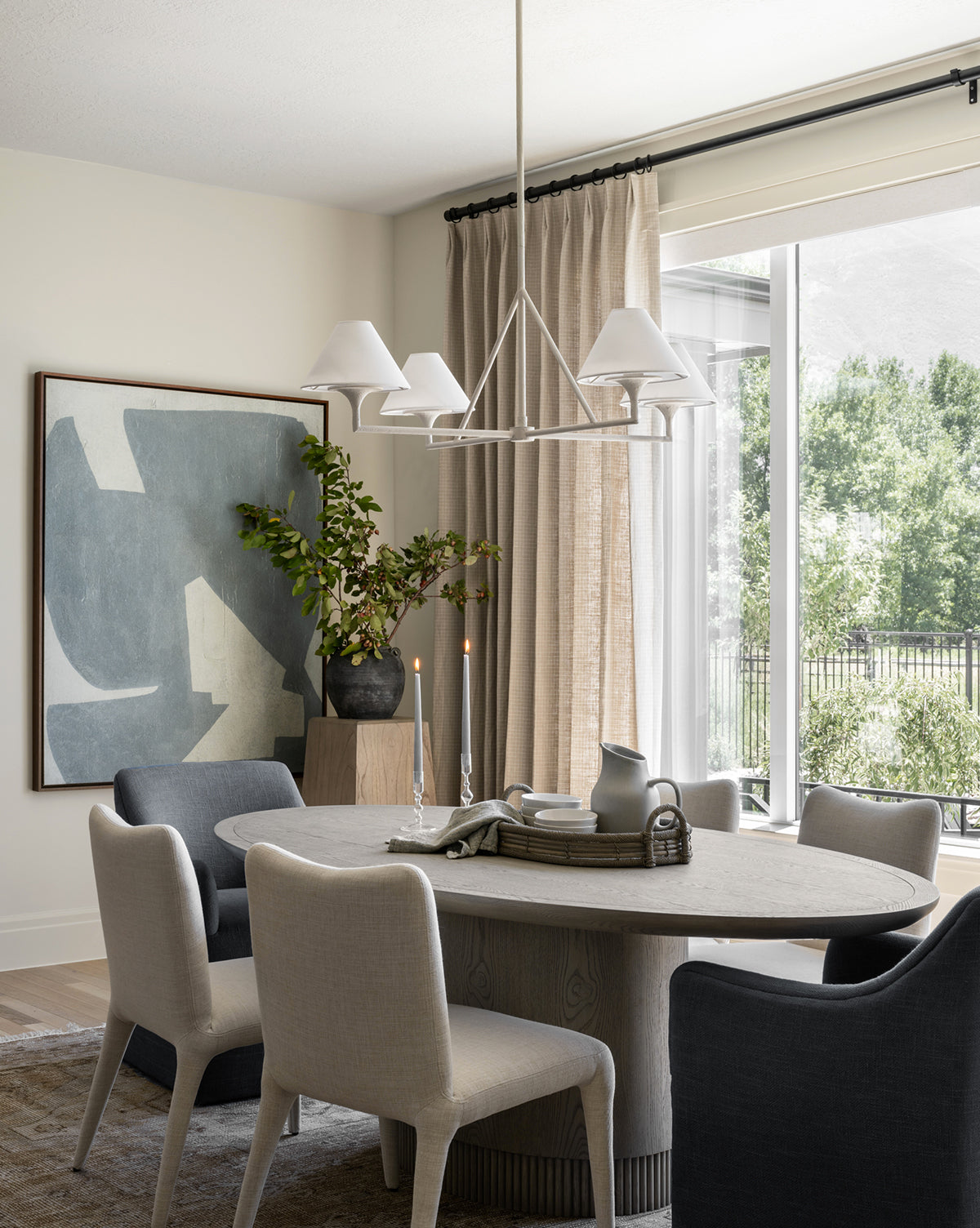 A modern dining room includes a round wooden table with six Elbert Dining Chairs in flax linen, neutral tones, an abstract painting, tall vase with greenery, candles, and large windows with beige curtains letting in natural light by McGee & Co.