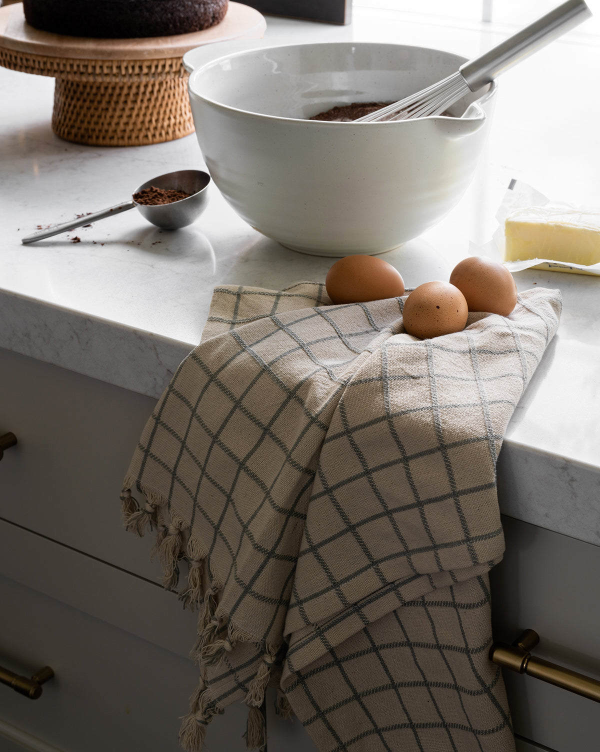 A kitchen counter features the Fullmer Stoneware Batter Bowl with a whisk, a checkered towel holding three eggs, a stick of butter, cocoa powder in a measuring spoon, and a chocolate cake on a stand in the background, by McGee & Co.