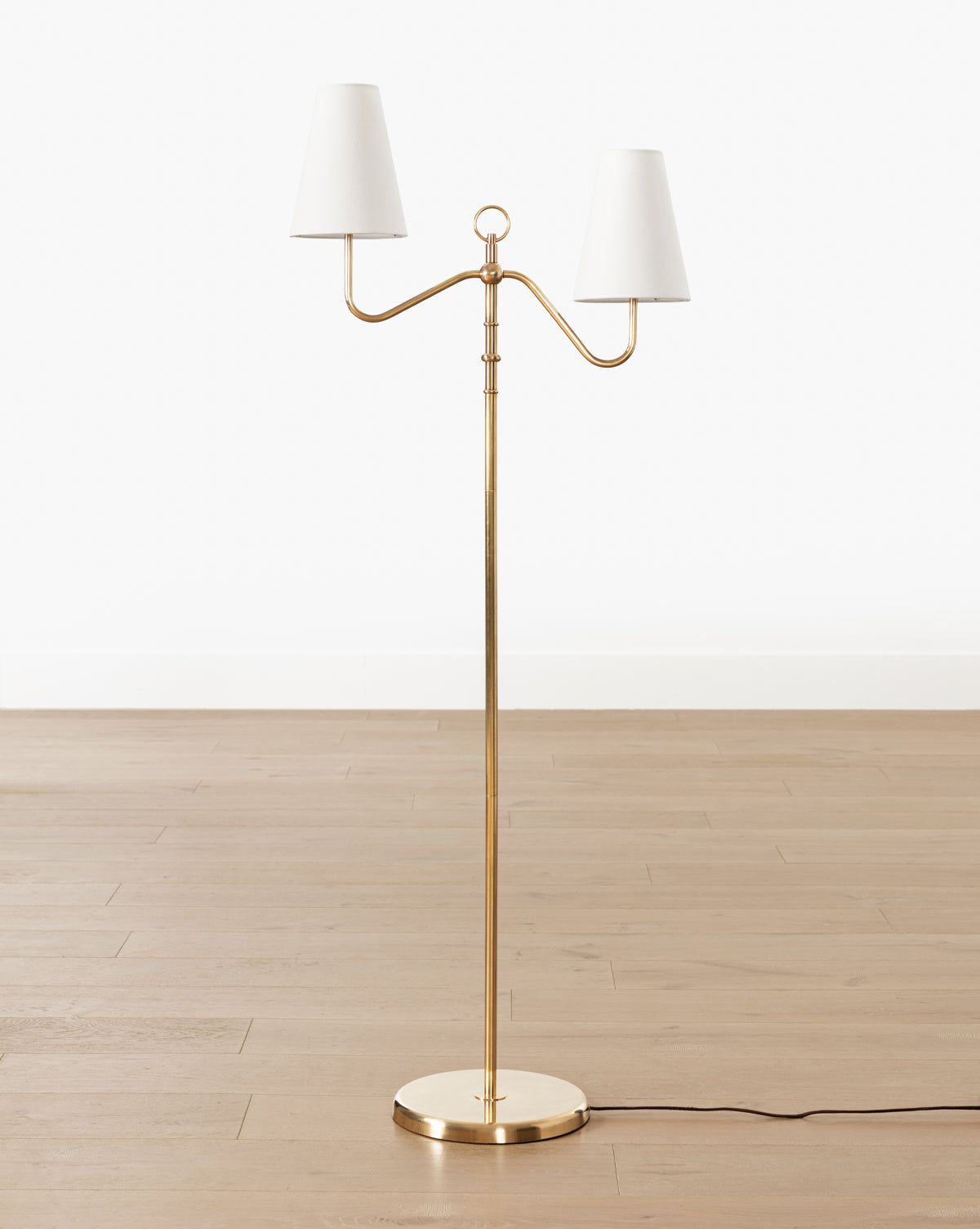 The Holloway Floor Lamp showcases an antique brass finish, double-arm design, two white lampshades, and a round base, complemented by a black electrical cord, by McGee & Co.