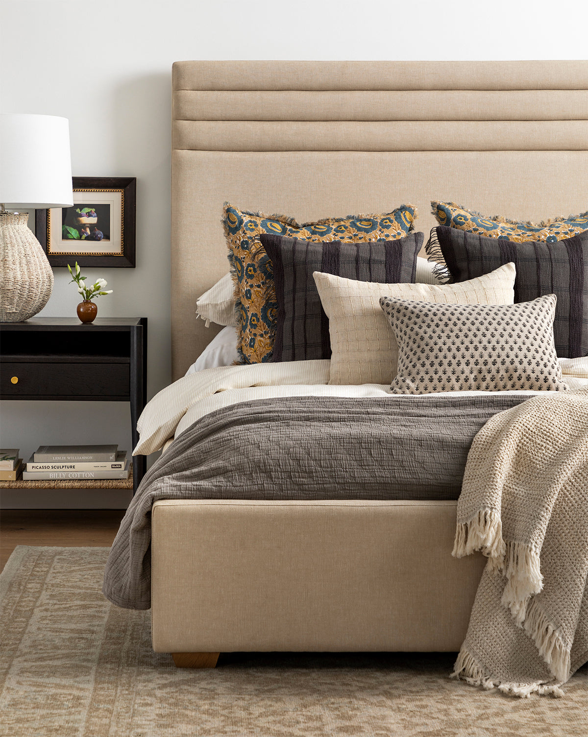 A beige upholstered bed with plush pillows, a textured blanket, and a taupe rug sits beside a black nightstand topped with a lamp, books, and plant. The wall features ‘Figs Still Life’ artwork for an elegant touch.