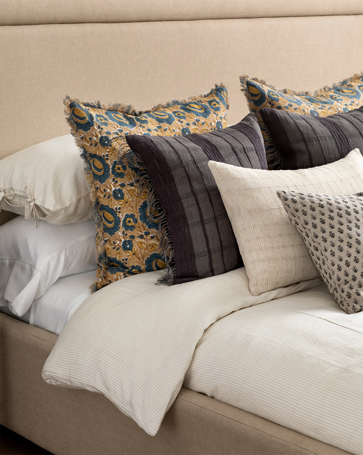 A neatly made bed with the Easton Striped Duvet Cover in cream and oatmeal, accented by layered decorative pillows in floral, plaid, and geometric patterns featuring blue, gold, gray, and cream tones.