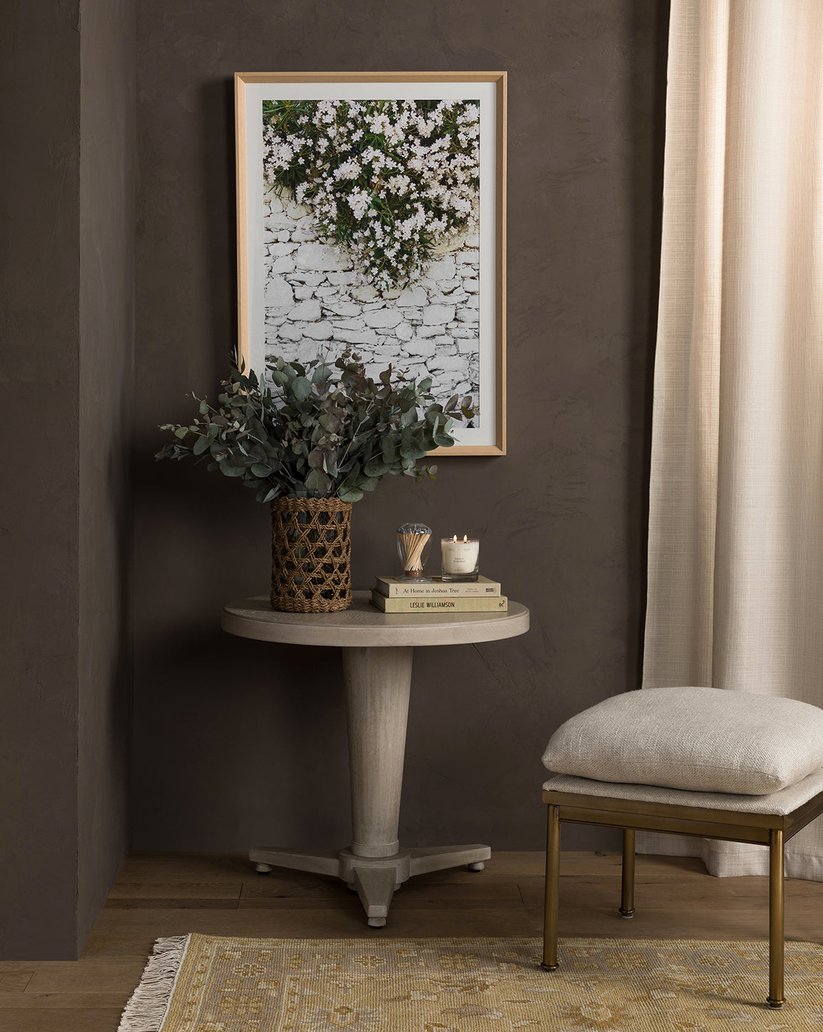 A small round table with decor sits in front of a dark wall, topped by the Stone & Blossoms botanical giclee print. Nearby are a cream curtain, an upholstered stool, and a light rug on the wood floor.