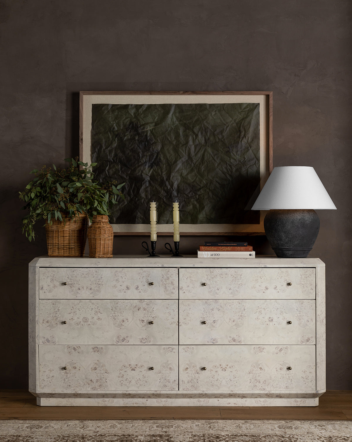 A cream-colored six-drawer dresser stands against a dark wall. On top are a black lamp with white shade, two candlesticks, stacked books, the Ahnamia Wicker Jar holding a leafy plant, and a large framed abstract artwork.